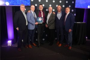 Six men in suits pose on a stage; the center man holds a 'WINNER' award plaque, with a purple-lit backdrop and circular logos behind them.