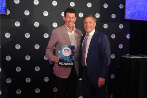 Two men in suits pose together at an awards ceremony; one holds a plaque that reads WINNER with a blue starry design and a circular event logo backdrop behind them.