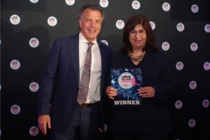 Two well-dressed people posing on a step-and-repeat backdrop; the woman holds a 'WINNER' award plaque for Long Island Choice Awards.