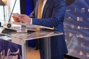 Man in a blue suit speaks at a clear podium with a microphone during a 'Top Lawyers' event backdrop.