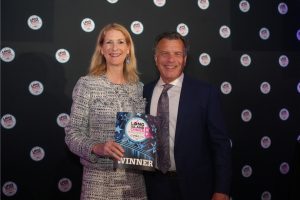 Two smiling adults in formal attire posing at an awards event, with the woman holding a 'WINNER' certificate against a backdrop of logos.