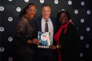 Three smiling attendees pose with a winner's plaque at an awards event, the man in a blue suit center flanked by two women in dark outfits.