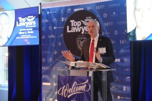 A man in a dark suit and red tie delivers a speech at a clear podium during a 'Top Lawyers of Long Island' event, with a blue branded backdrop behind him and a welcome banner in front of the podium.
