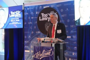 Man in a suit giving a speech at a clear podium, with a blue 'Top Lawyers' backdrop and a 'Welcome' sign on the lectern.