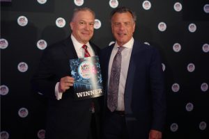 Two men in suits posing at an awards event; the left man holds a 'WINNER' certificate with a blue star design.