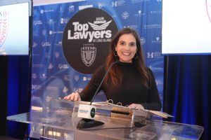 Woman in a black top smiles while speaking at a clear podium at a 'Top Lawyers of Long Island' event with a blue sponsor backdrop behind her.