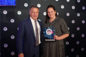 Man in a dark blue suit and a woman in a patterned dress pose together at an awards event; the woman holds a 'WINNER' certificate.