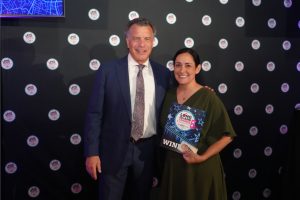 Man in a navy suit and a woman in a green dress pose for a photo at an awards backdrop dotted with circular logos, the woman holding a WIN certificate.