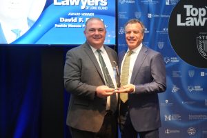Two men in dark business suits pose with a glass trophy at a law awards event, standing in front of a blue sponsor backdrop.