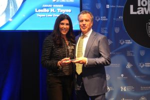 Woman and man in business attire pose on a stage, holding an award trophy that reads 'Top Lawyers' with Leslie H. Tayne mentioned on the display background.