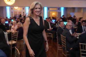 Smiling blonde woman in a black sleeveless dress walks through a banquet hall filled with seated guests at a formal event.