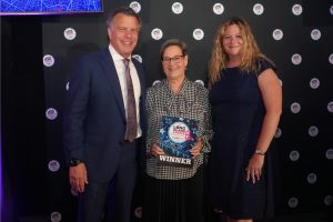Three people posing together at an awards event; the central woman holds a WINNER plaque, flanked by a man in a suit and a woman in a dark dress, with a logo-filled backdrop.