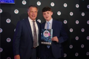 Two men in dark suits posing on a stage; the younger man holds a 'WINNER' award plaque at the Long Island Choice Awards.