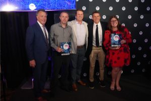 Five people on a stage at an awards event; two winners hold plaques reading 'WINNER' while the others smile.
