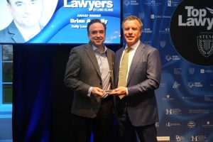 Two men in suits hold a glass award and pose for a photo on a stage at a lawyers’ awards event, with a blue backdrop and “Top Lawyers” branding.