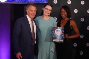 Three people pose on a stage at an awards ceremony: a man in a dark suit, a smiling woman in a sparkly teal dress, and a second woman in black holding a WINNER certificate against a backdrop of event branding.