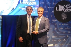 Two men in suits smiling on stage, holding a glass award, with a blue backdrop that reads 'Top Lawyers of Long Island'.