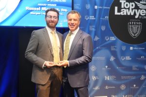 Two men in suits pose with a glass award on stage at a legal awards ceremony, blue backdrop filled with sponsor logos behind them.