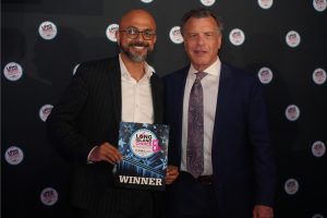 Two men in suits pose at an awards event; the man on the left holds a blue WINNER plaque with Long Island Choice Awards branding in front of a branded backdrop.