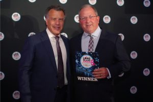 Two men in dark suits stand together at an awards event, one holding a 'WINNER' plaque with the Long Island Choice Awards logo. A backdrop covered with circular logos is visible behind them.
