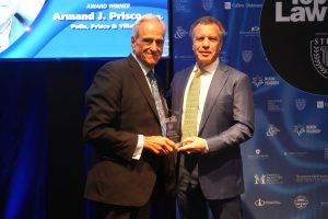Two men in suits on a stage, one holding a glass trophy, during an awards ceremony with a blue sponsor backdrop and a screen that reads 'Award Winner'.