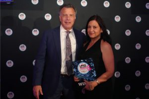 Man in a navy suit and a woman in a black dress stand together on a dark backdrop covered with circular logos; the woman holds a blue "Long Island Choice Awards" plaque, smiling at the camera.