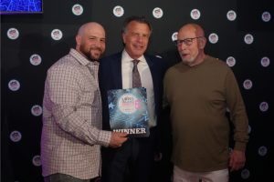 Three men posing at an awards event; the center man in a suit holds a 'WINNER' plaque against a dark backdrop with circular logos on it.