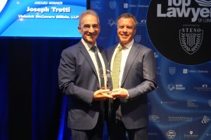 Two smiling men in dark suits on stage, each holding a glass award together at an awards ceremony.