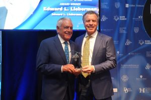 Two men in dark suits smiling while presenting a glass award at a formal event. Behind them is a blue backdrop with sponsor logos.