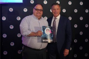 Two men pose with a winner certificate at an awards event; backdrop features circular logos on a dark wall.