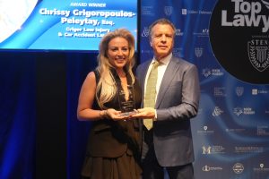 Smiling woman in a black dress and a man in a suit pose with a glass award on a stage; screen behind reads 'Chrissy Grigoropoulos-Peleyat, Esq.'