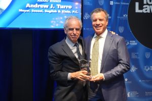 Two men in dark suits pose with a glass trophy on stage at a lawyers awards event, smiling in front of a blue sponsor backdrop.