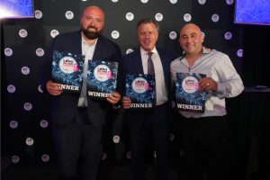 Three men in suits smiling while holding Long Island Choice Awards winner plaques against a dark backdrop.