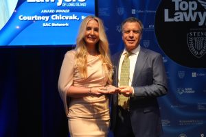 A woman in a pale pink dress and a man in a navy suit pose on a stage, holding a glass award together against a blue backdrop.