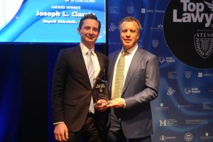 Two men in suits pose on stage, one receiving a glass 'Top Lawyers' award trophy.