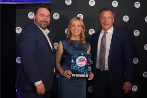 Three formally dressed people pose on stage; center woman holds a 'Winner' plaque at the Long Island Choice Awards backdrop