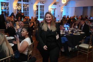 Woman in a black suit walks through a formal banquet hall, smiling, as guests dine at round tables in the background.
