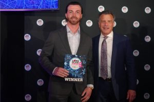 Two men in suits pose on stage; the younger holds a blue “WINNER” award plaque with starry design, smiling at the camera.