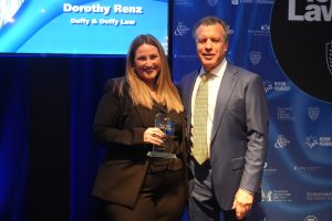 Woman in a black suit holds a glass award on stage beside a man in a blue suit at an awards event; screen behind reads ‘Dorothy Renz’.