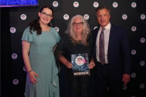 Three adults posing for a photo at an awards event; center woman holds a WINNER plaque with a blue starry design, flanked by a woman in a teal dress and a man in a suit.