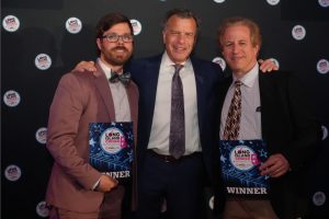 Three men in suits pose together at an awards night, two holding Long Island Choice Awards winner plaques against a dark backdrop.