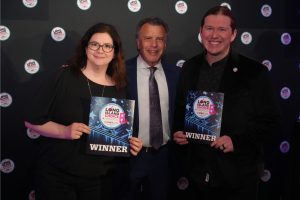 Three people pose with award certificates at a dark backdrop; two winers hold 'WINNER' plaques while a suited man stands between them.