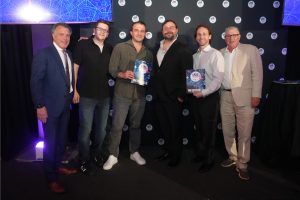 Six men pose on a stage at an awards event; two hold 'WINNER' certificates, with a backdrop of circular logos.