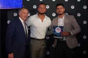 Three men posing on a dark backdrop at an awards event; the man on the right holds a WINNER plaque with a blue star design.