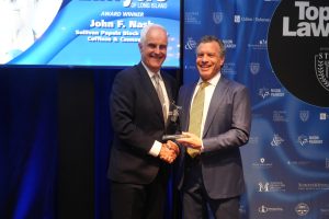 Two men in dark suits shake hands on a stage; one holds a glass award while a blue sponsor backdrop reads 'Top Lawyers' and 'John F. Nash' in the background.