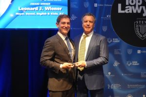 Two men in dark suits pose on stage, one presenting a glass award to the other, with a blue sponsor backdrop and the words 'Award Winner' visible on a screen behind them.