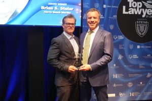 Two men in suits posing on stage with a glass award; screen behind reads 'Award Winner Brian S. Stolar Harris Beach Murtha'.