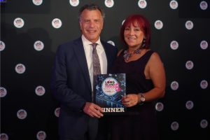 Man in a navy suit and a woman in a dark purple dress smile for the camera while holding a blue and white award plaque that says 'WINNER' at a Long Island Choice Awards event, backdrop covered with circular logos.