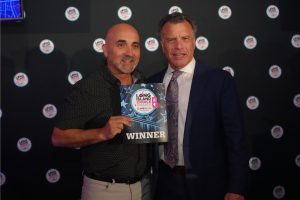 Two men pose together at an awards event; one holds a sign reading WINNER with a trophy graphic behind him and a dark backdrop dotted with circular logos.