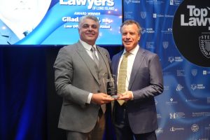 Two suited men smile while presenting a glass award on stage at a lawyers awards event, with a blue backdrop and 'Top Lawyers' branding behind them.